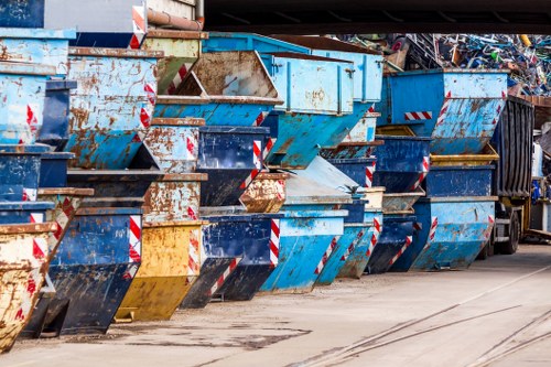 A row of large metal skips or dumpsters lined up outside on a paved surface, with some showing signs of rust and paint chipping in various shades of blue, yellow, and black. The skips are stacked closely together, with their open tops visible, and are positioned parallel to a building or structure in the background. To the right, part of a large waste removal truck can be seen, likely used for collecting and transporting rubbish from the skips. The environment suggests an industrial or commercial area, possibly related to waste management services in Canary Wharf or surrounding London districts, used by companies like Office Clearance Canary Wharf for rubbish removal. The surface appears clean but shows some dark stains consistent with typical waste handling zones, and the lighting is natural, indicating daytime conditions with overcast or partly cloudy weather. The scene is typical of a designated rubbish disposal site prepared for waste collection, highlighting the importance of proper skip and rubbish management in urban environments.