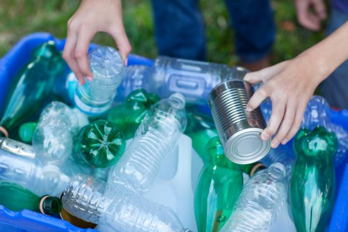 A close-up view of a person's hands sorting recyclable household waste outdoors, with one hand holding a clear plastic water bottle and the other placing a metal tin into a recycling bin. The bin contains various empty plastic bottles, mainly clear and green, with some crushed or partially flattened. The background includes a grassy area with blurred greenery and the edge of a person's denim-clad leg visible. The image emphasizes the importance of proper rubbish sorting and recycling, aligning with waste management services such as those provided by Office Clearance Canary Wharf, in a typical outdoor setting in London’s Docklands area. The lighting is natural, suggesting daylight, and the scene highlights the textures of the plastic bottles and metallic cans, underscoring environmentally conscious rubbish disposal practices.
