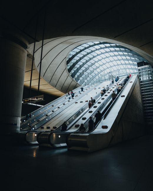 Interior view of a modern transportation hub featuring multiple escalators arranged in parallel, leading upwards beneath a curved glass ceiling that allows natural light to illuminate the space. The e