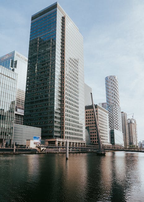 A black and white photograph of a modern city skyline along a river, featuring multiple high-rise buildings with varying architectural styles. The tallest building is a rectangular glass tower with a