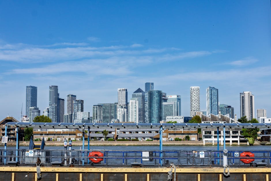 A panoramic view of a city skyline during dusk, featuring a mix of modern high-rise office buildings and residential structures along a riverbank. The skyscrapers display various architectural styles,