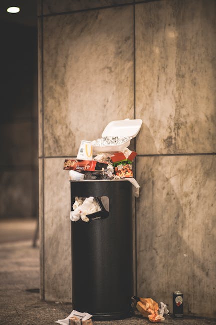 A human hand extending from the right side of the image is dropping a crumpled piece of brown paper into a white plastic recycling bin labeled 'PAPER' in bold black letters. The bin is positioned on a