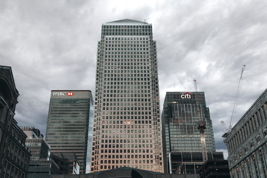 A panoramic view of a modern cityscape at dusk, featuring several tall office buildings with glass facades reflecting the evening sky. The buildings include a mixture of rectangular and curved archite