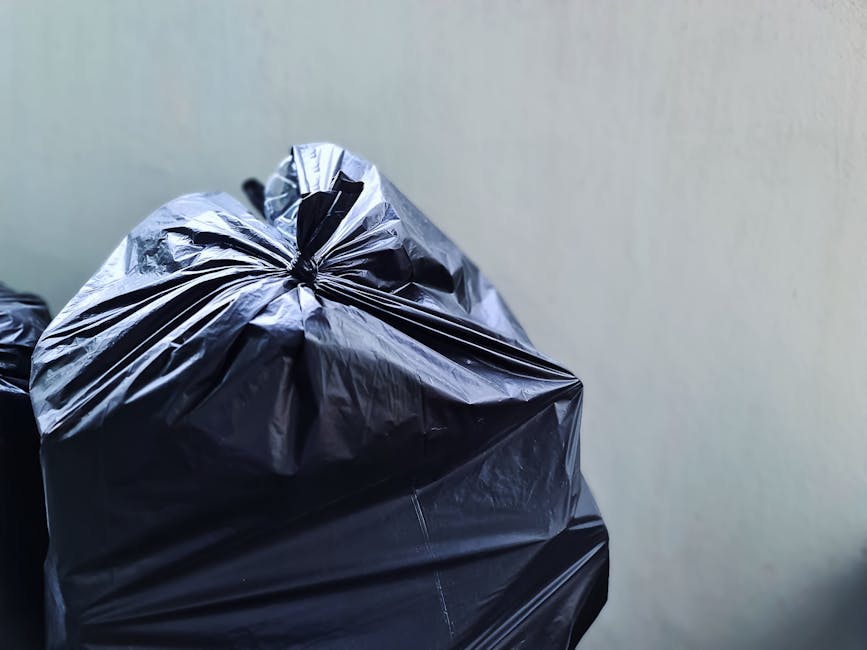 Close-up image of a black plastic rubbish bag with a slightly glossy surface, tightly sealed at the top with a gathered, twisted neck. The bag is filled with waste materials, giving it a rounded, bulg